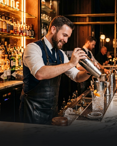 Professional bartender preparing a cocktail behind a stylish bar counter, pouring a mixed drink from a shaker into a glass in a warm, upscale lounge setting with liquor bottles displayed in the background.
